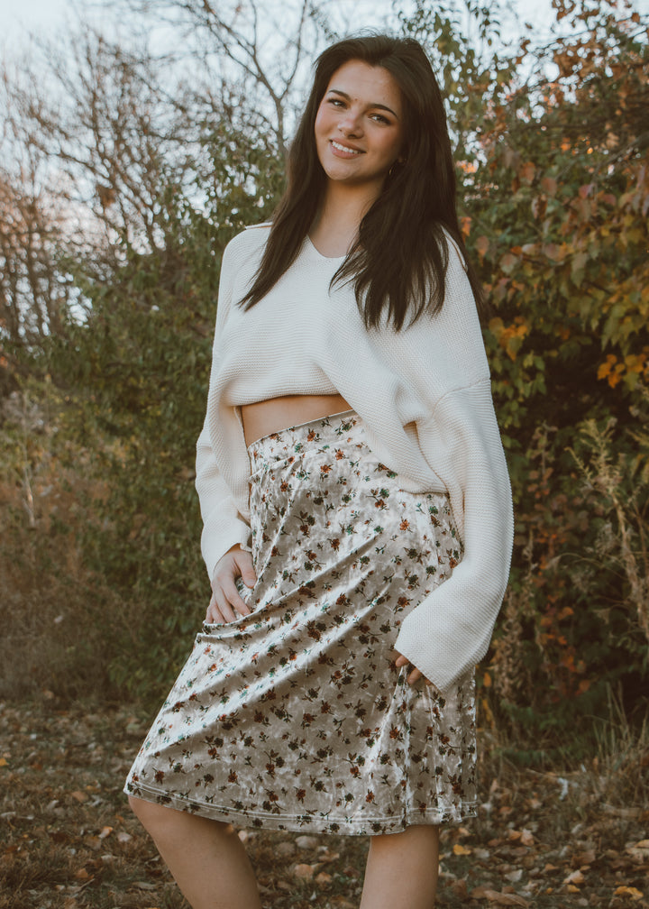 Woman wearing a white sweater and patterned skirt standing outdoors with trees in the background