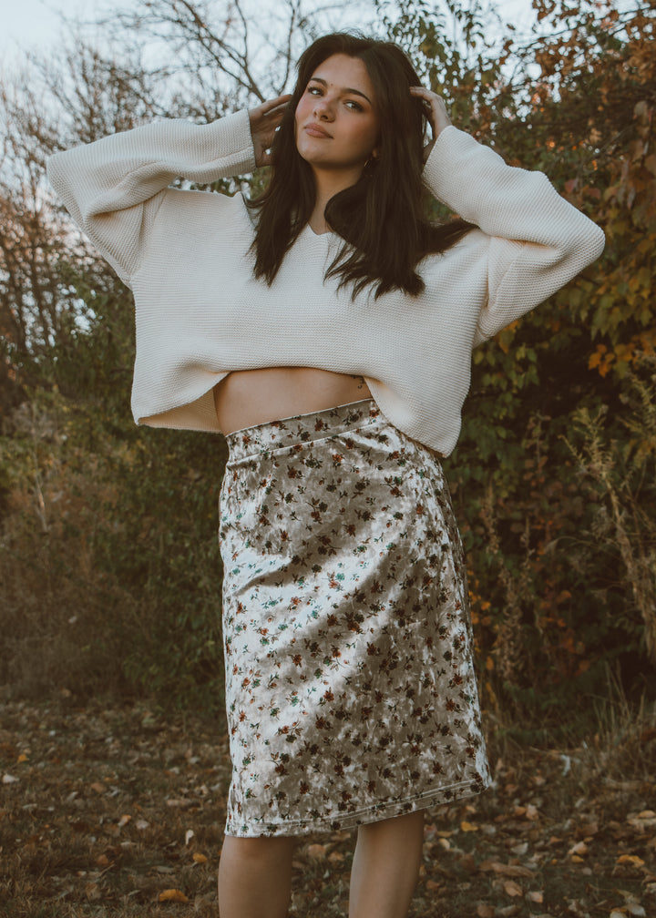 Woman wearing a white sweater and floral skirt standing outdoors with trees in the background