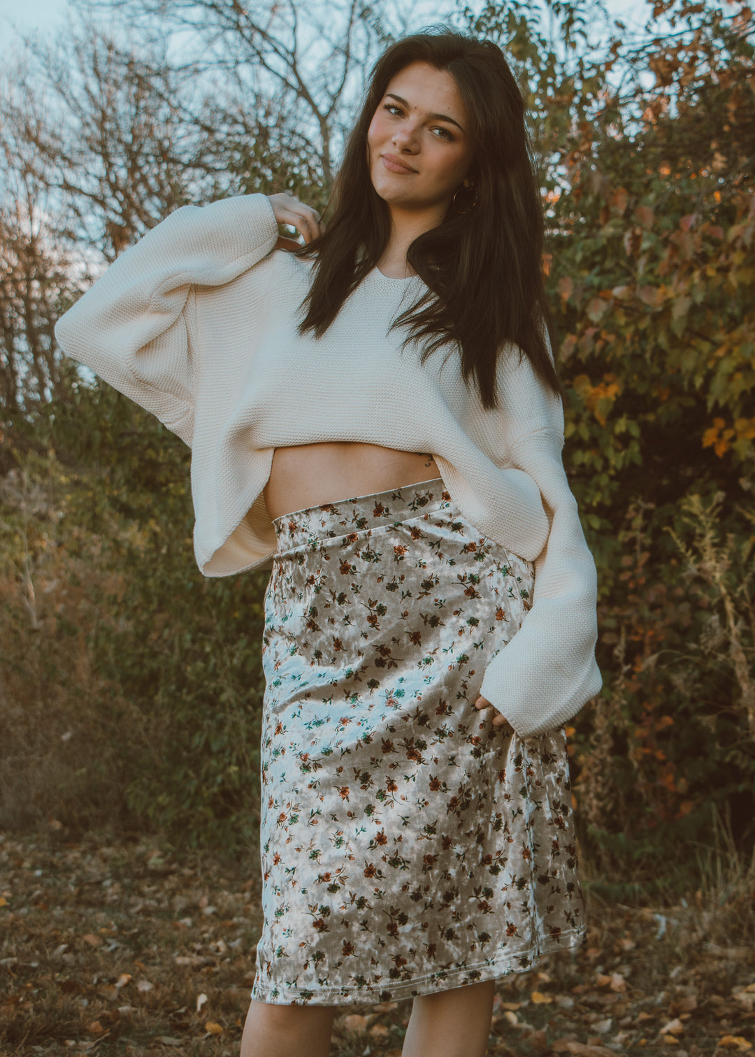 Woman wearing a white sweater and floral skirt standing outdoors with trees in the background
