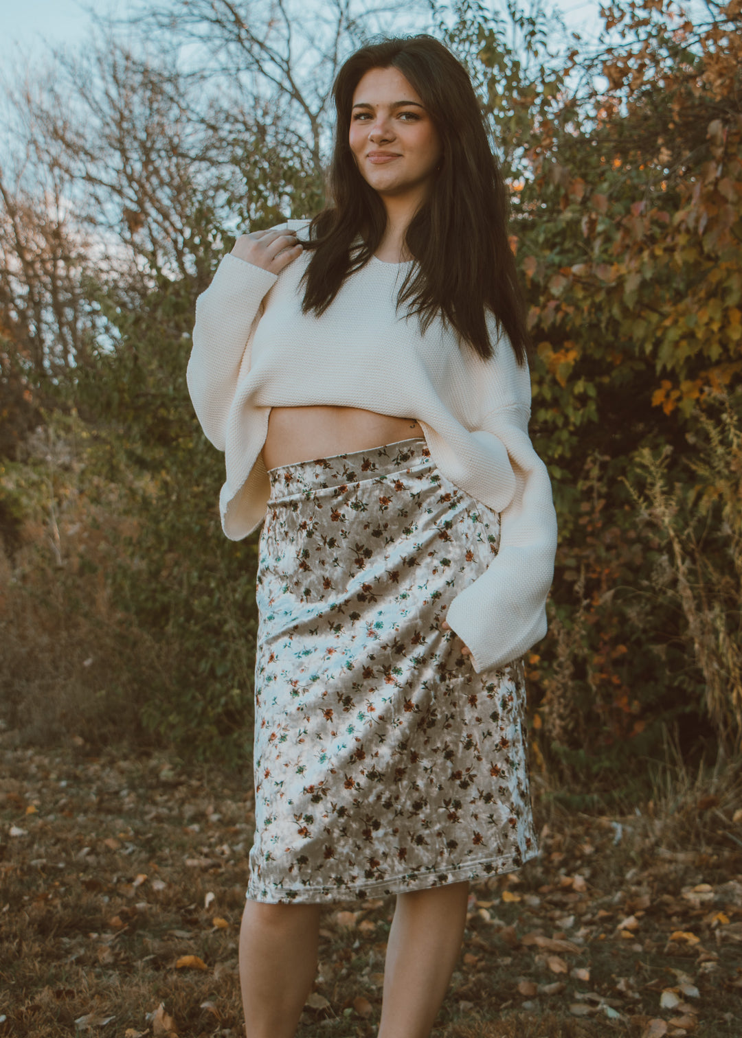 Woman wearing a white sweater and floral skirt standing outdoors with trees in the background