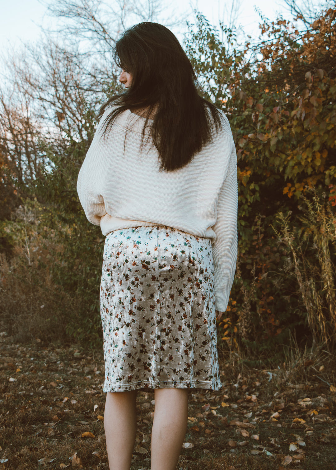 Woman in a white sweater and floral skirt standing in a forest