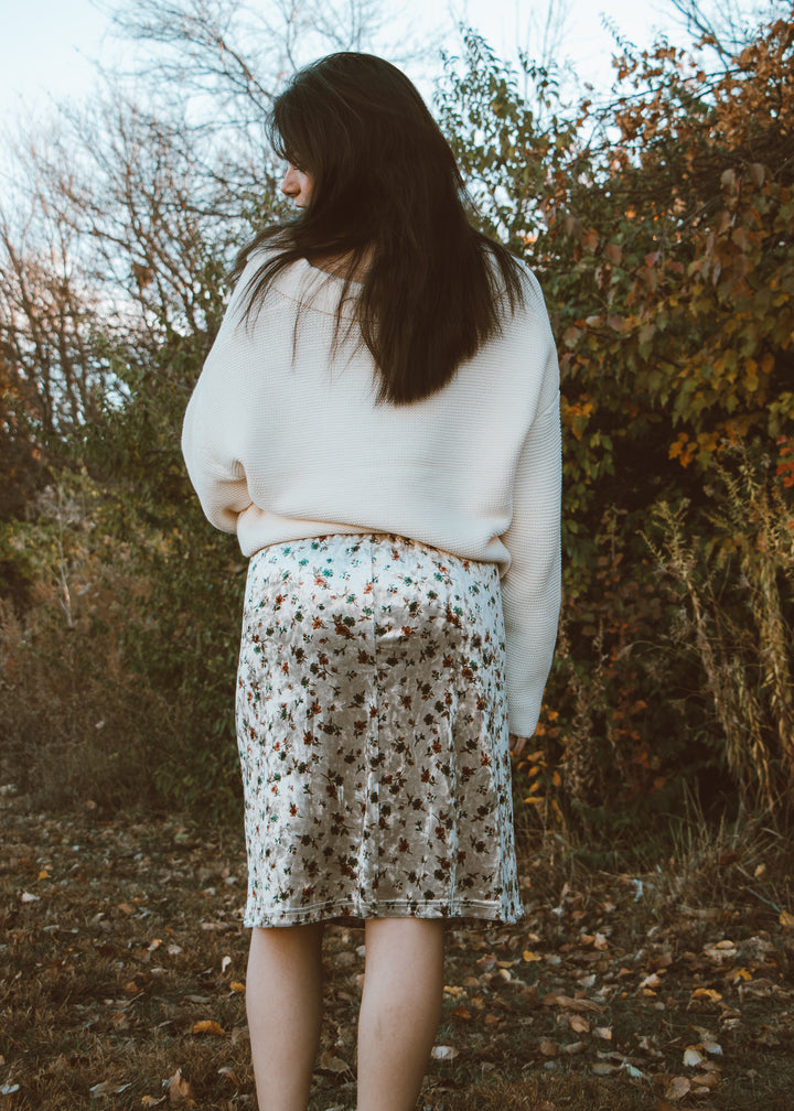 Woman in a white sweater and floral skirt standing in a forest