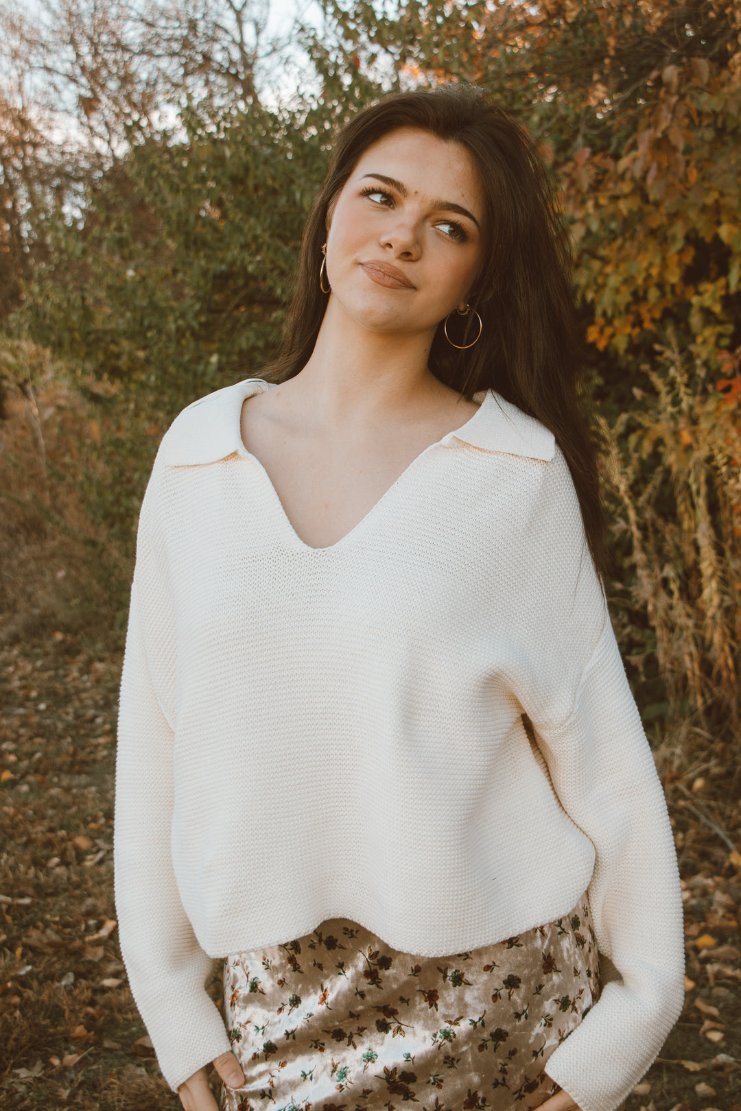 Woman wearing a white sweater and patterned pants standing in a natural setting.