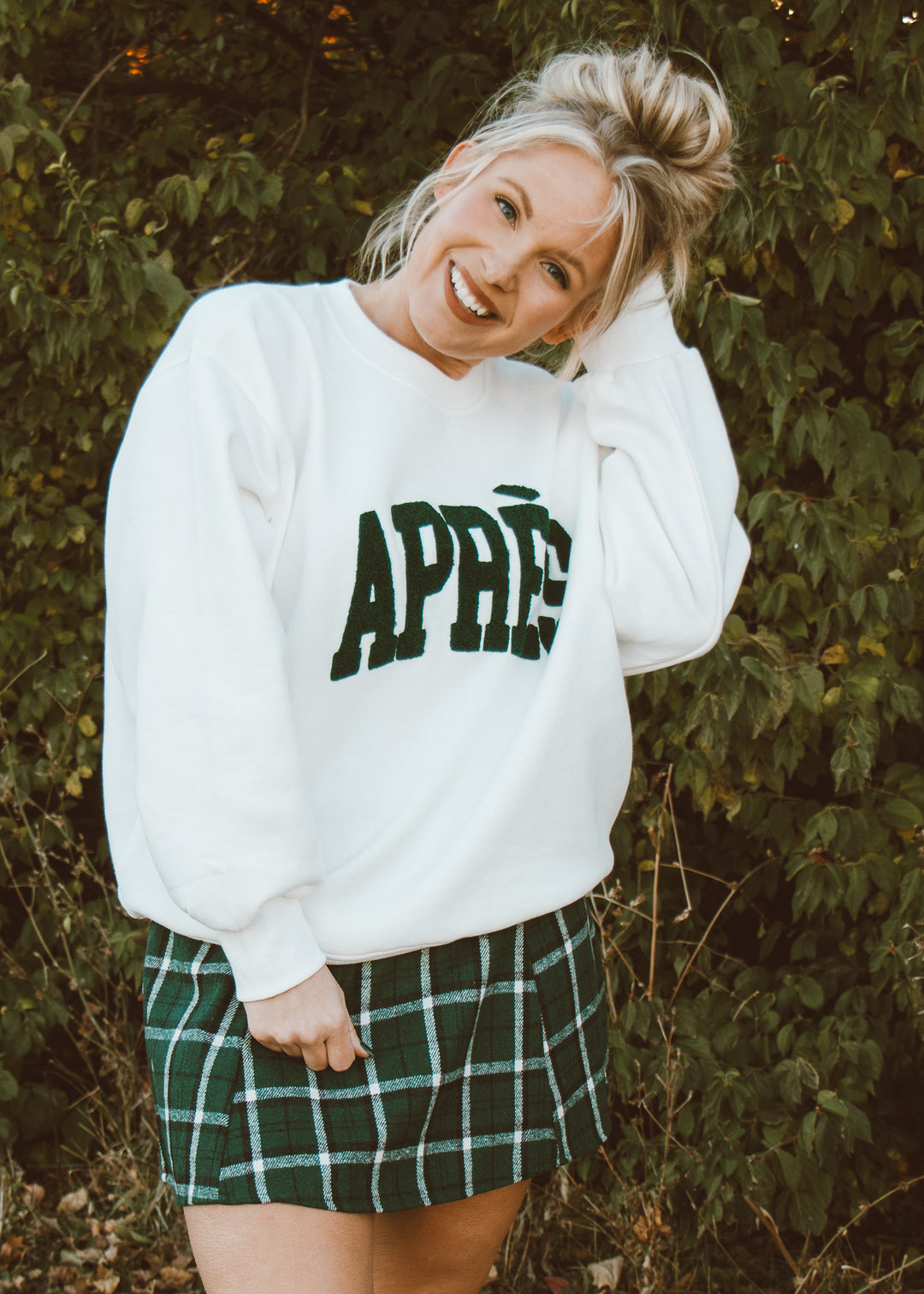 Person wearing a white sweatshirt with 'APRÈS' text and a green plaid skirt outdoors.