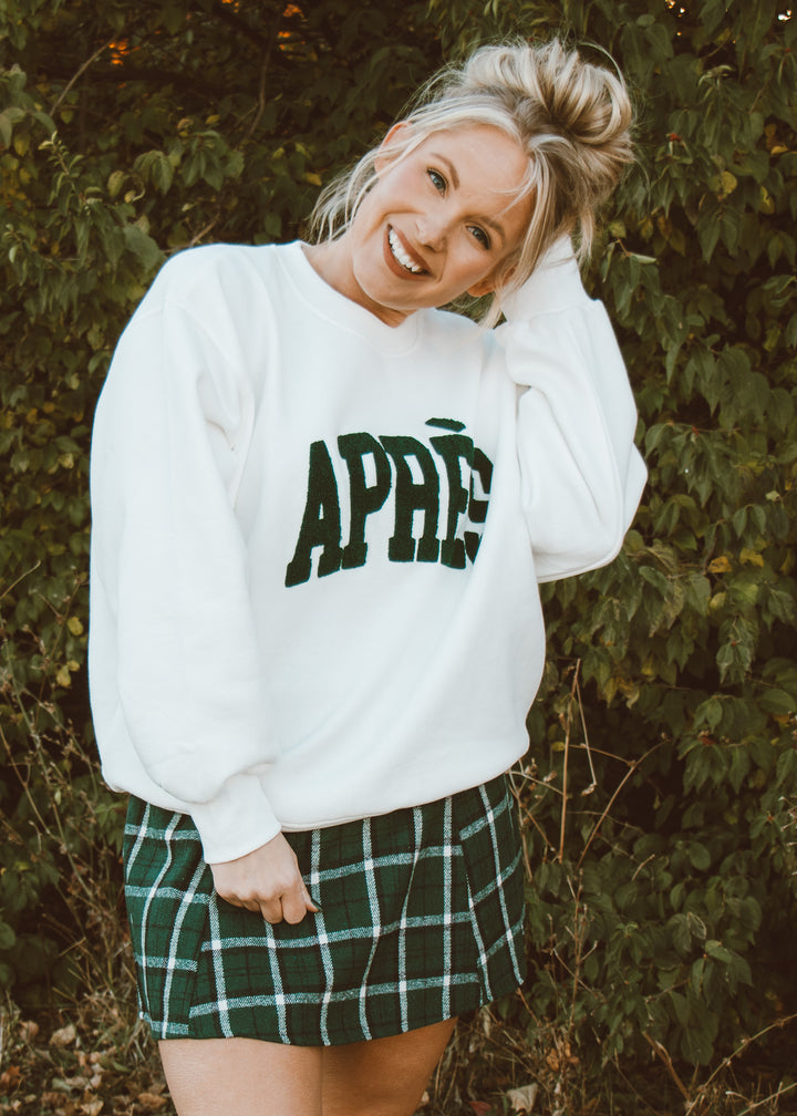 Person wearing a white sweatshirt with 'APRÈS' text and a green plaid skirt outdoors.