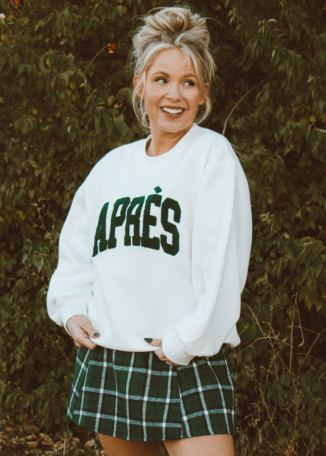 Woman wearing a white sweatshirt with 'APRÈS' text and a green plaid skirt outdoors.