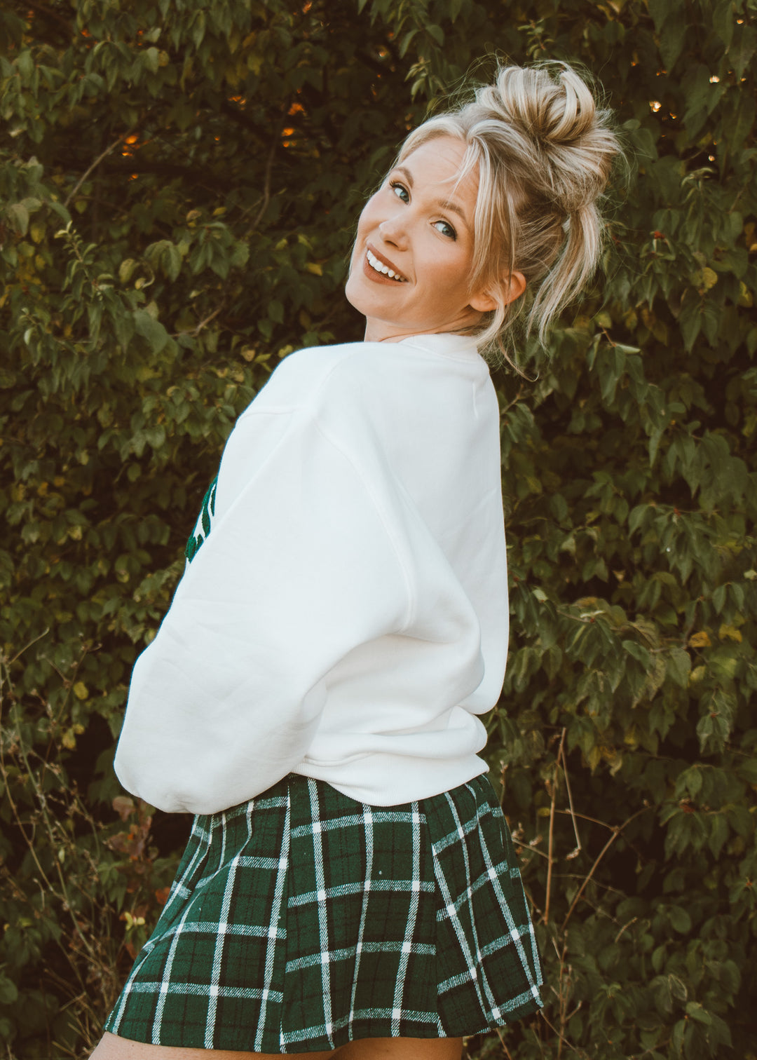 Woman wearing a white sweater and green plaid skirt standing against a leafy background