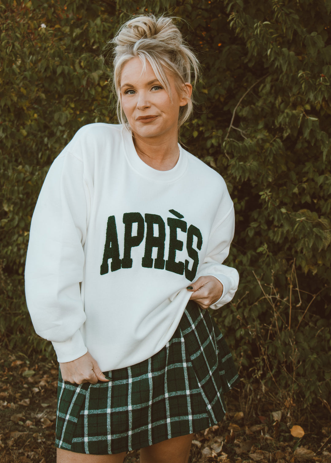 Woman wearing a white sweatshirt with 'APRèS' text and a plaid skirt outdoors.