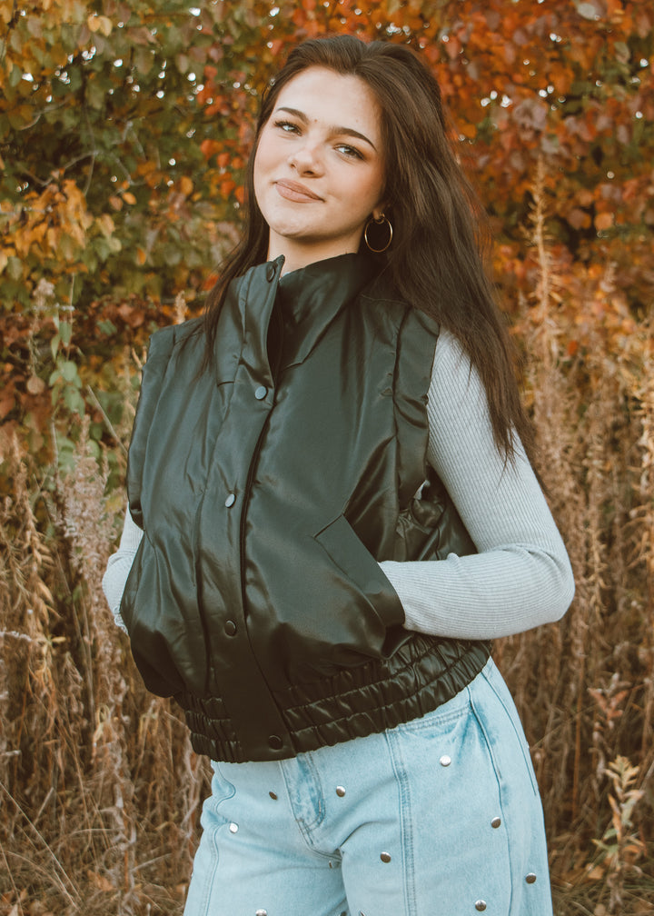 Woman wearing a green vest and light blue jeans standing in an autumn setting.