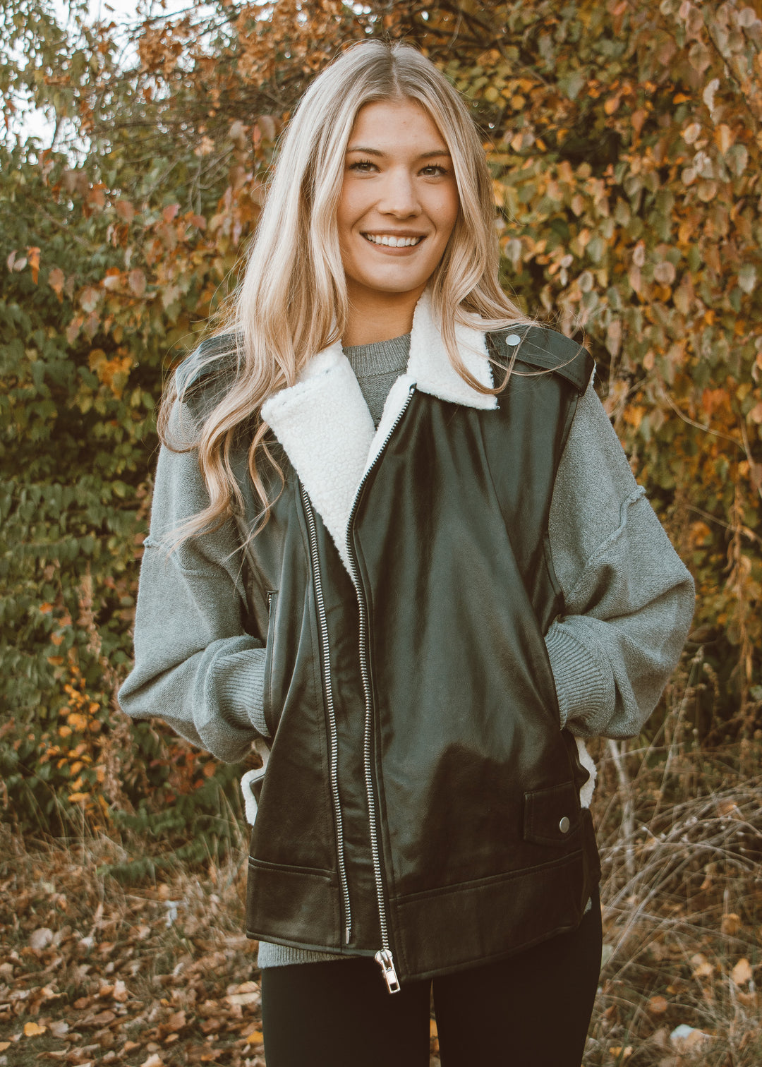 Woman wearing a green jacket with white fur lining, standing in an outdoor setting with trees in the background.