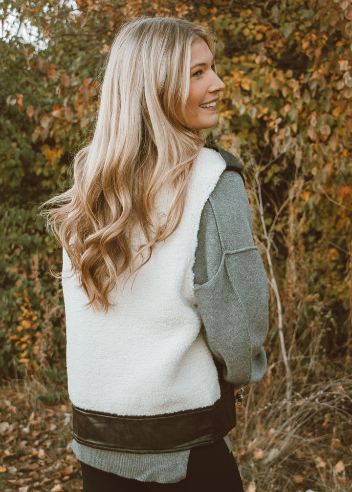 Woman wearing a white fleece vest over a green sweater in an outdoor setting with trees and leaves.
