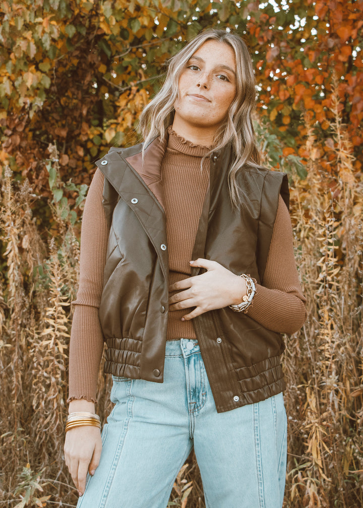 Woman wearing a brown vest over a brown sweater with light blue jeans, standing in front of autumn foliage.