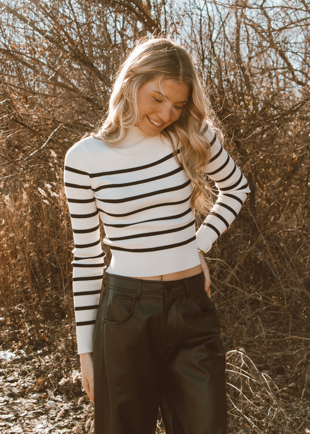 Woman wearing a black and white striped sweater and dark pants standing in a natural setting.