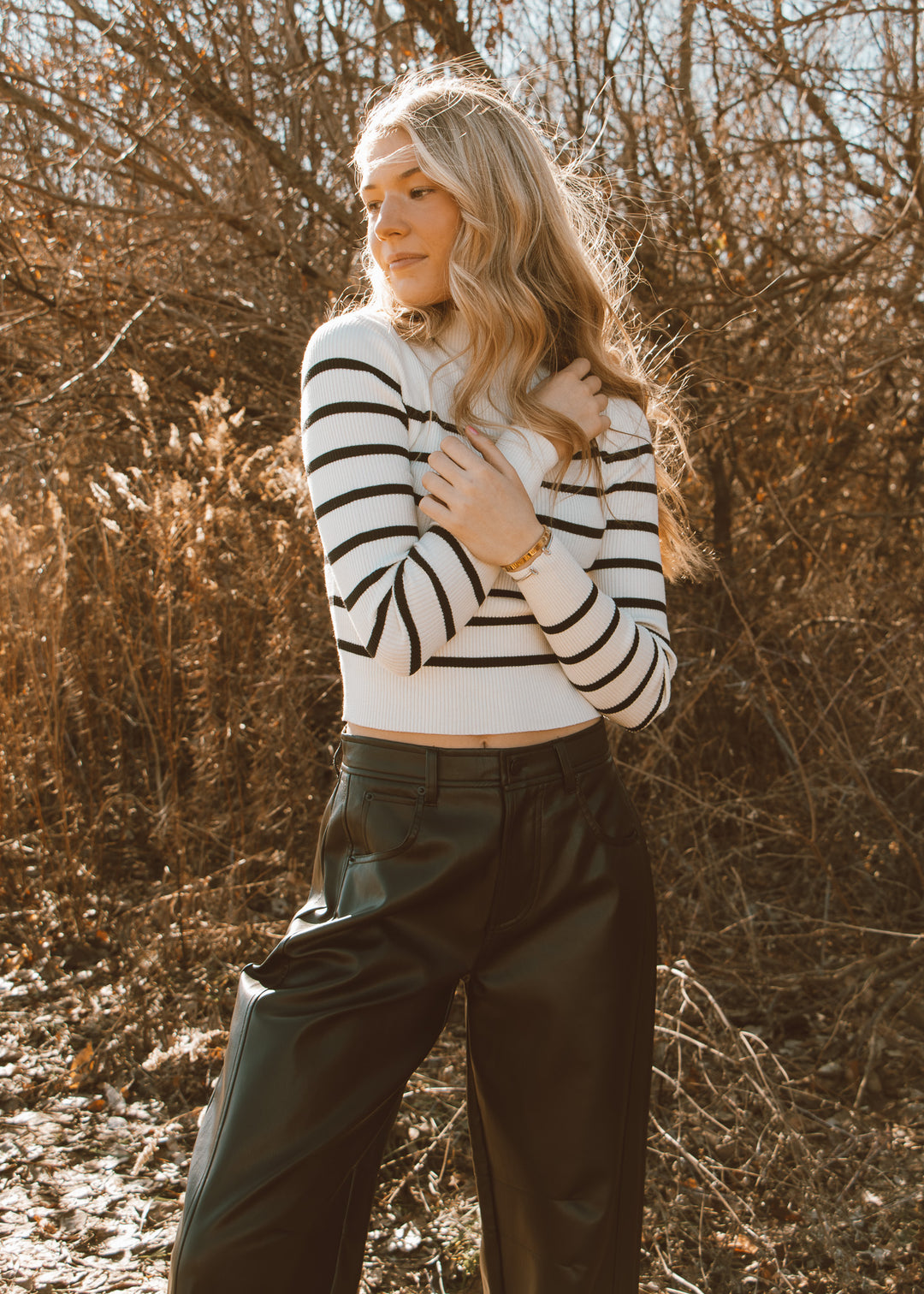 Woman wearing a black and white striped sweater and green pants standing in a natural setting with bare trees.
