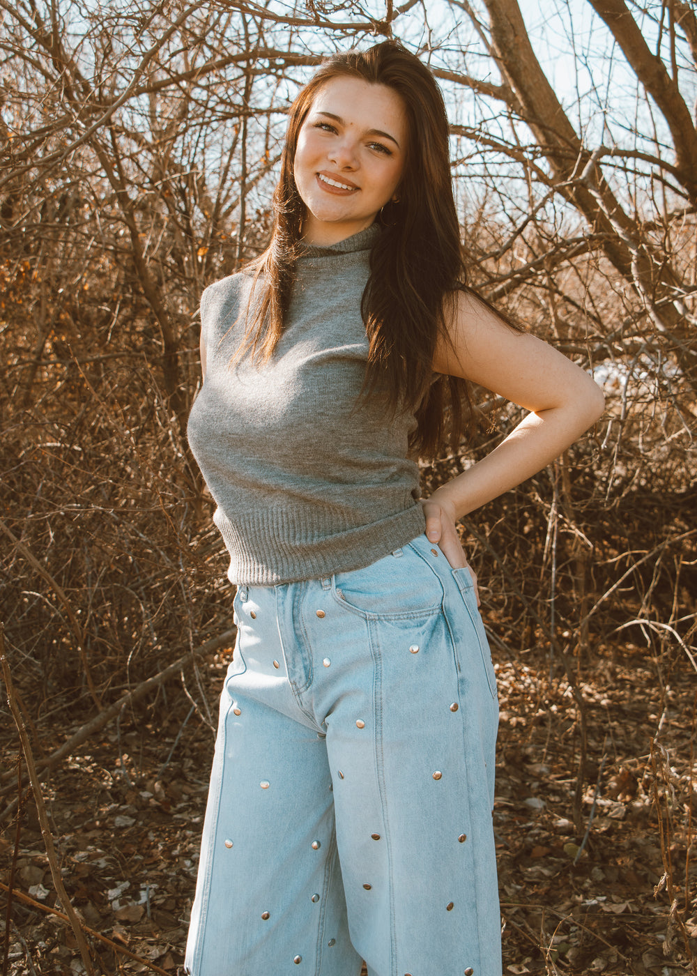 Woman wearing a gray sweater and light blue jeans standing in a natural setting with bare trees.