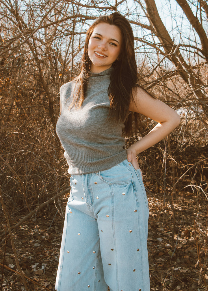 Woman wearing a gray sweater and light blue jeans standing in a natural setting with bare trees.