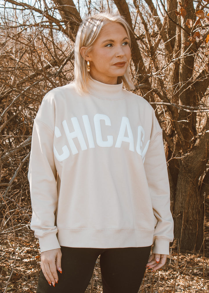 Woman wearing a beige sweatshirt with 'CHICAGO' printed on it, standing in front of leafless trees.