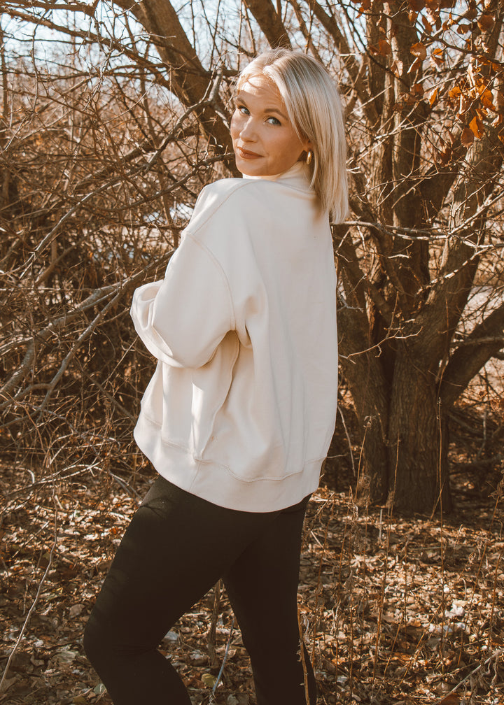 Person wearing a white sweater and black pants standing in a wooded area with bare trees.