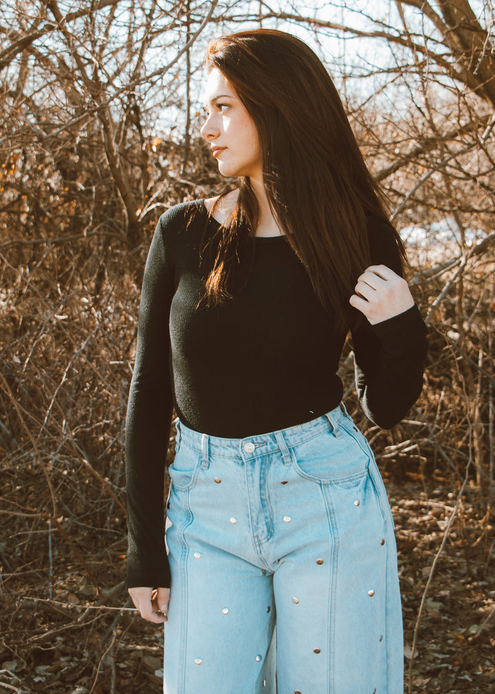 Woman wearing a black long-sleeve top and light blue jeans standing in a natural setting with bare trees.
