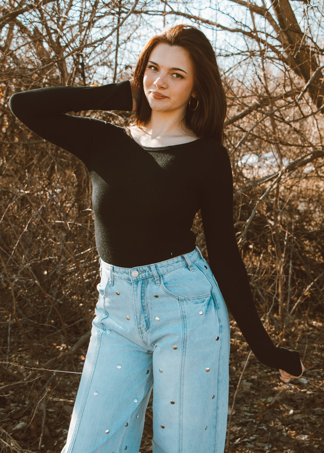 Woman wearing a black long-sleeve top and light blue jeans standing in a natural setting with bare trees.