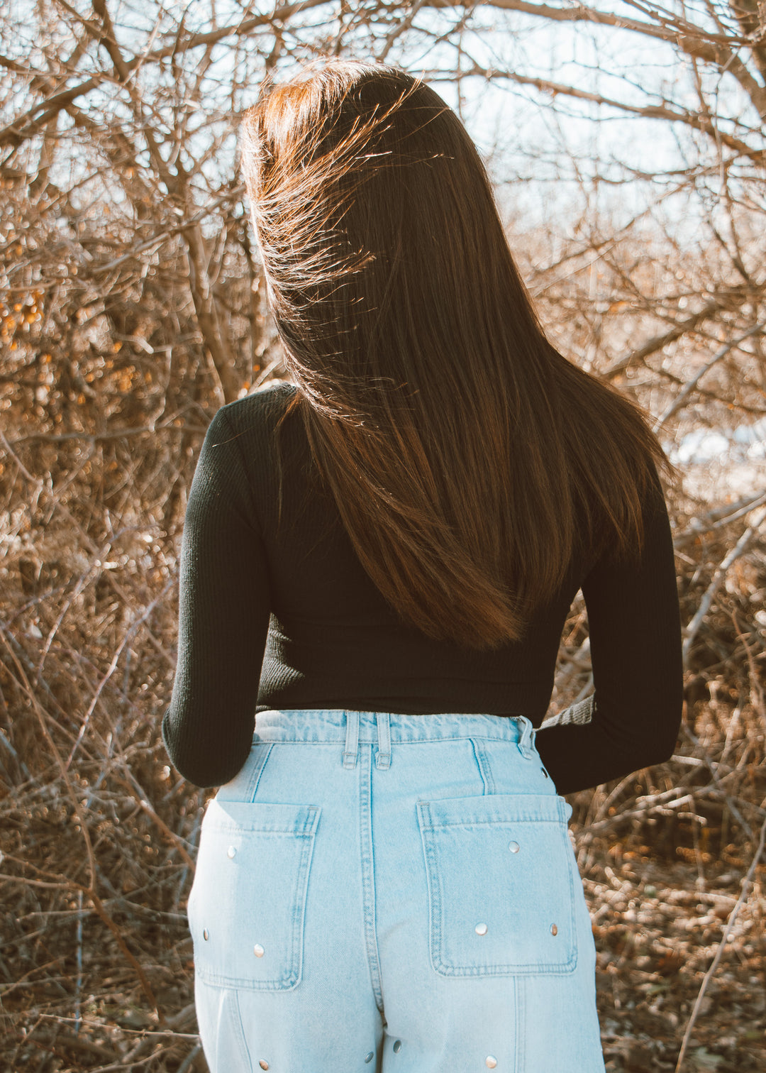 Person with long hair standing in a natural setting with bare trees.