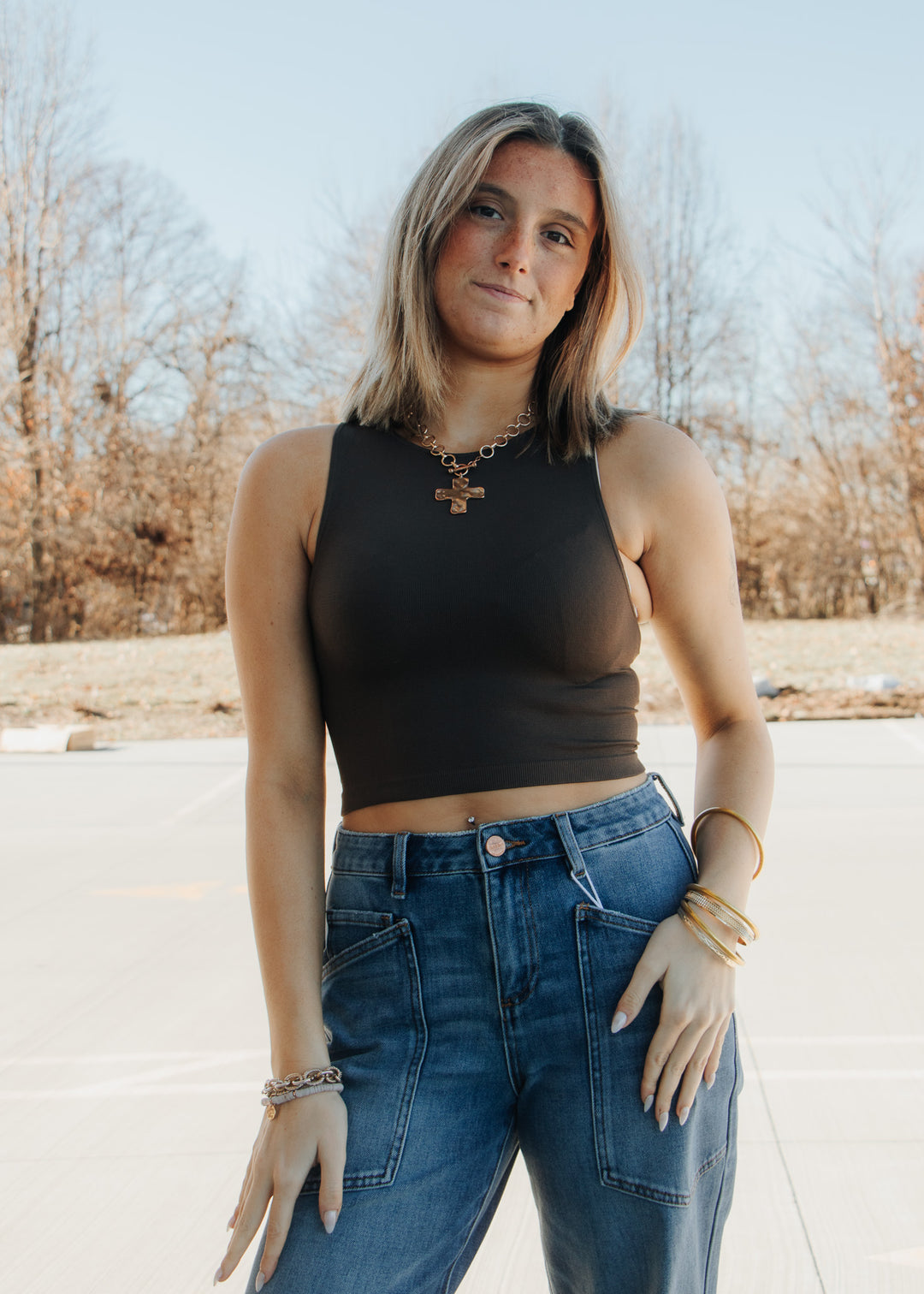 Woman wearing a brown sleeveless top and blue jeans standing outdoors with trees in the background.