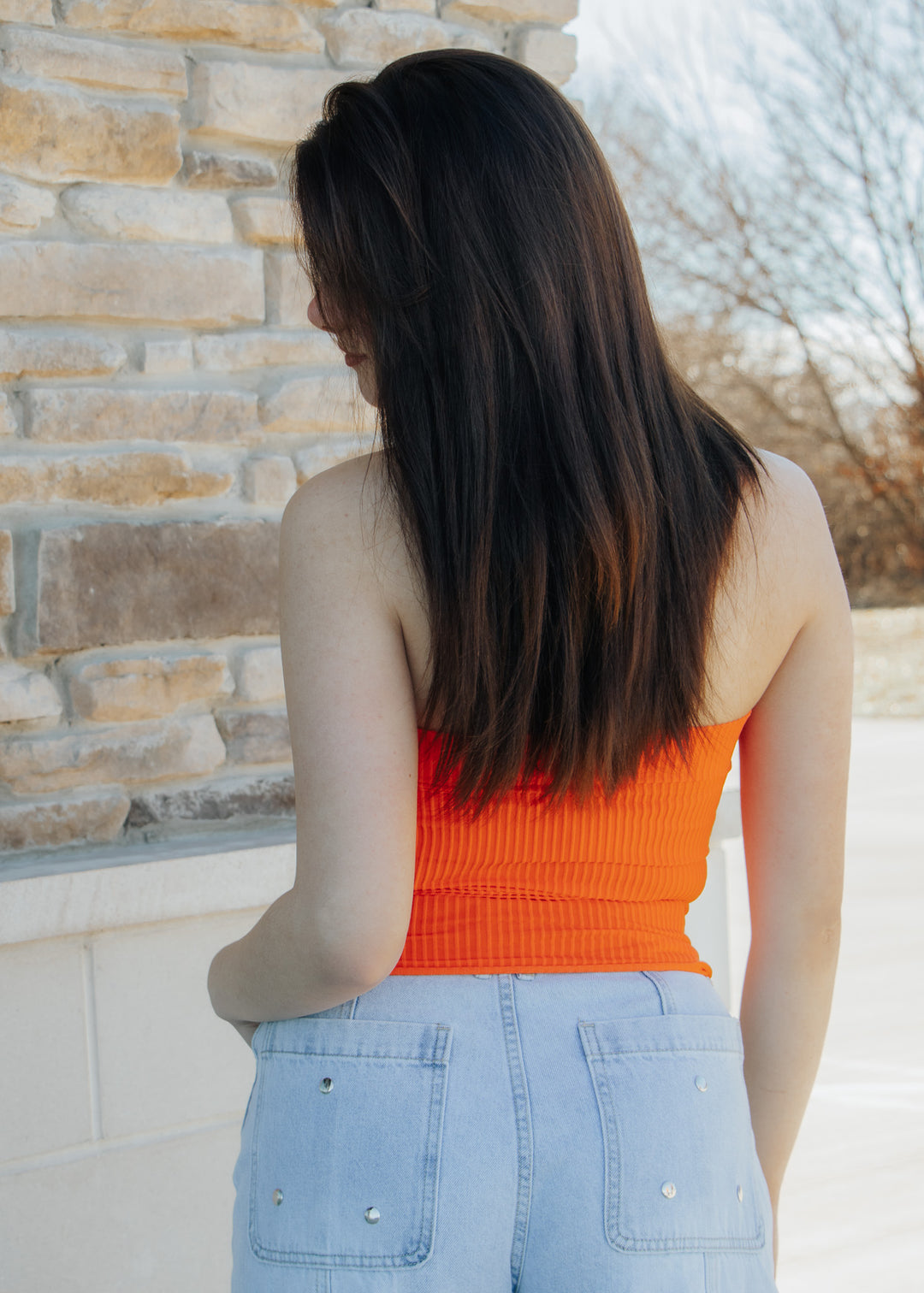 Person wearing an orange top and light blue jeans standing against a stone wall.