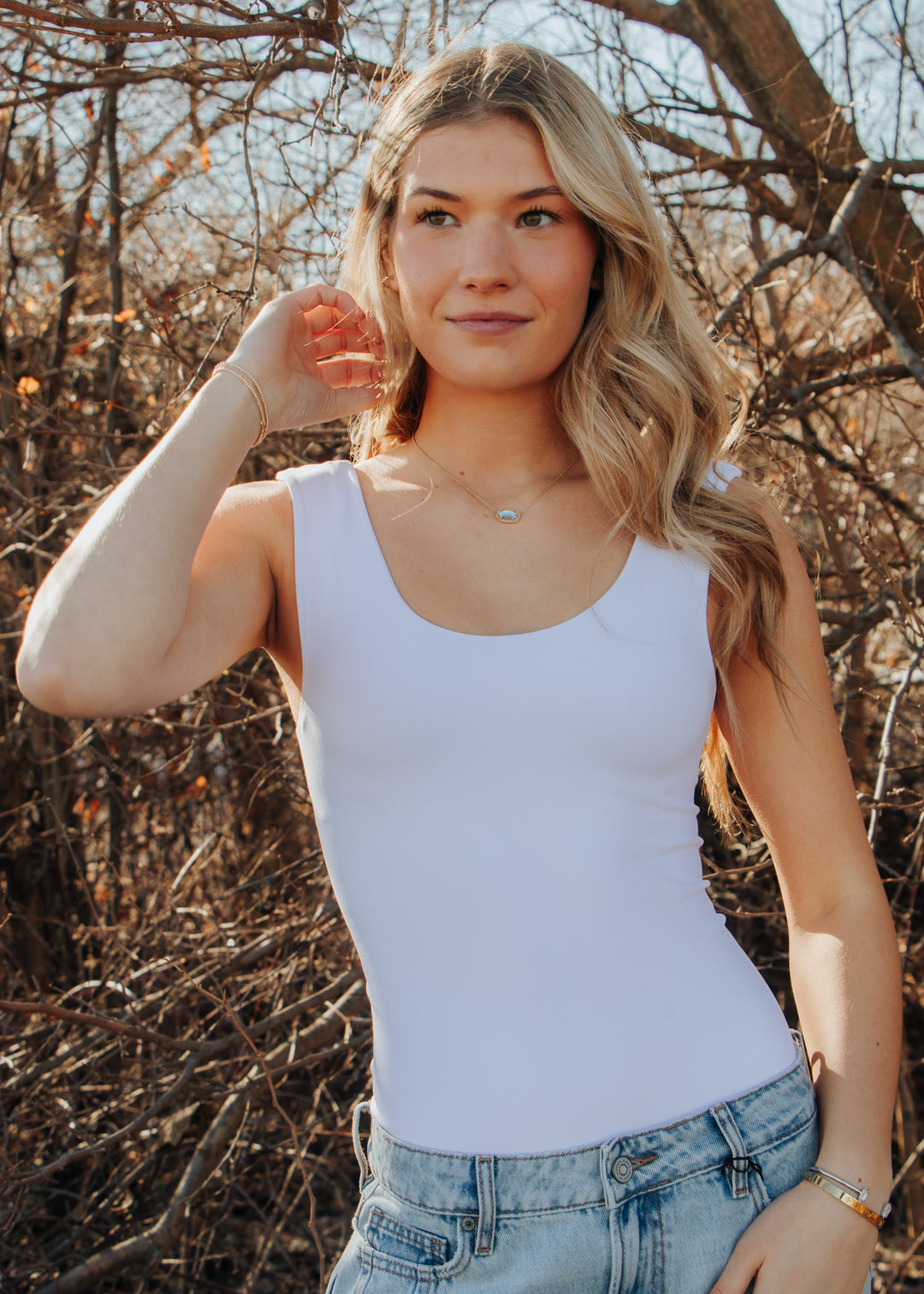 Woman wearing a white tank top and jeans standing in a natural setting with trees.