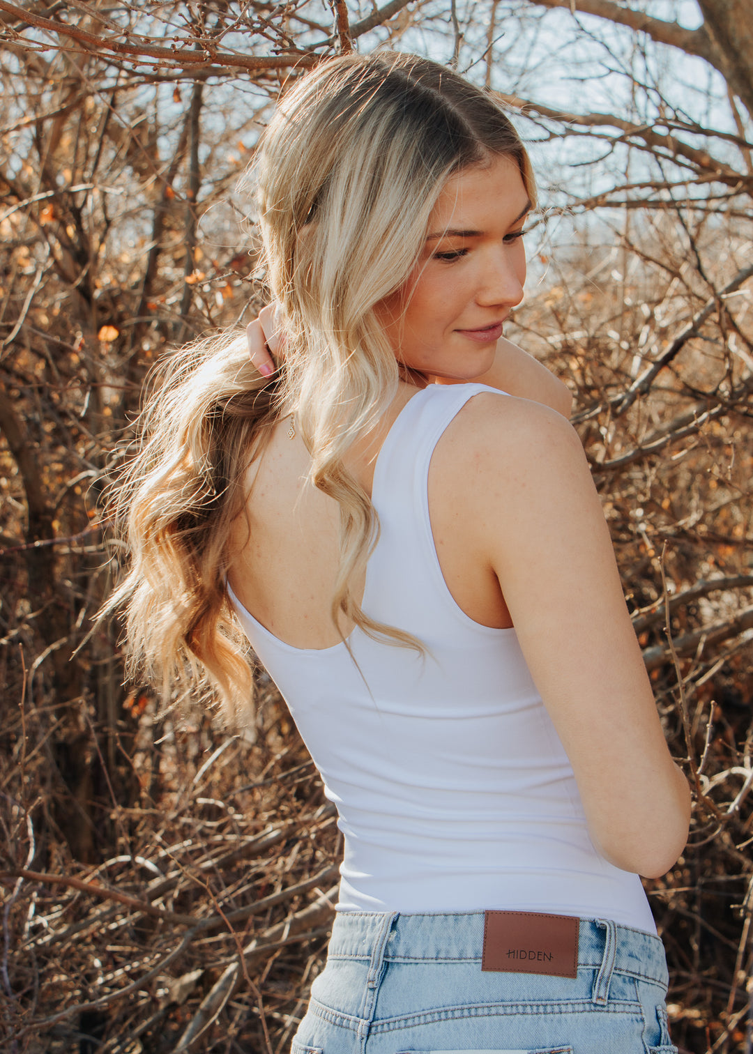 Woman in a white tank top and jeans standing among bare trees.