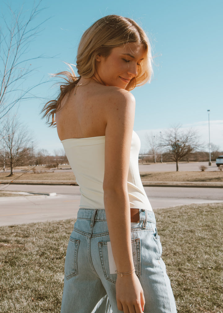 Woman wearing a white strapless top and light blue jeans standing outdoors on a sunny day.