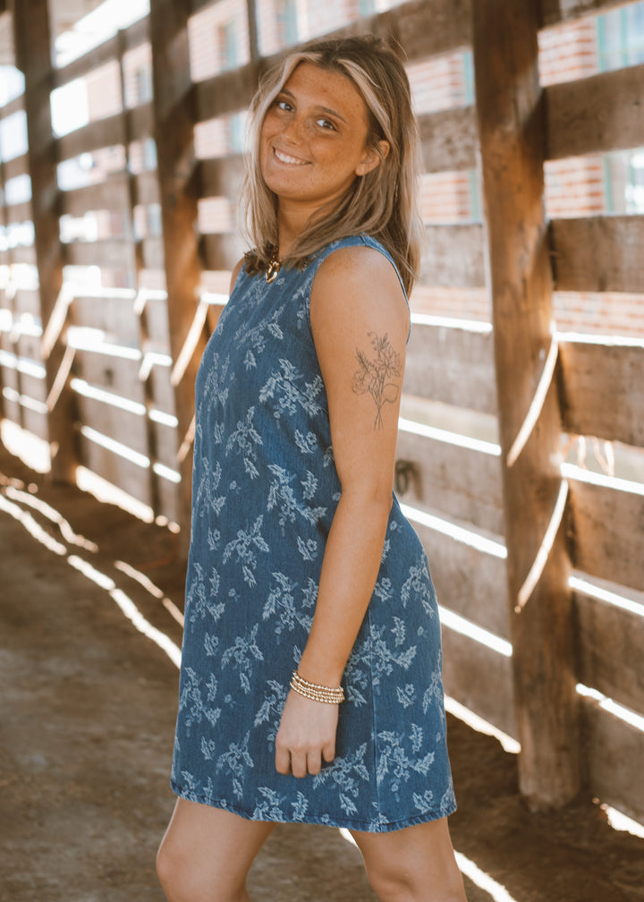 Woman wearing a blue dress with a floral pattern standing in front of wooden slats.