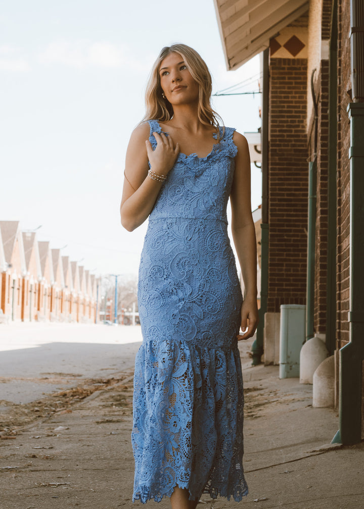 Woman in a blue lace dress standing outdoors near a building.
