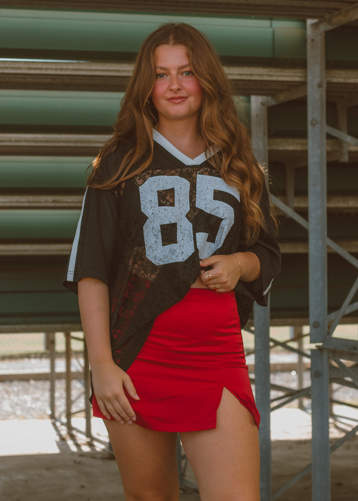 Woman wearing a sports jersey with the number 85 and a red skirt in an outdoor setting.