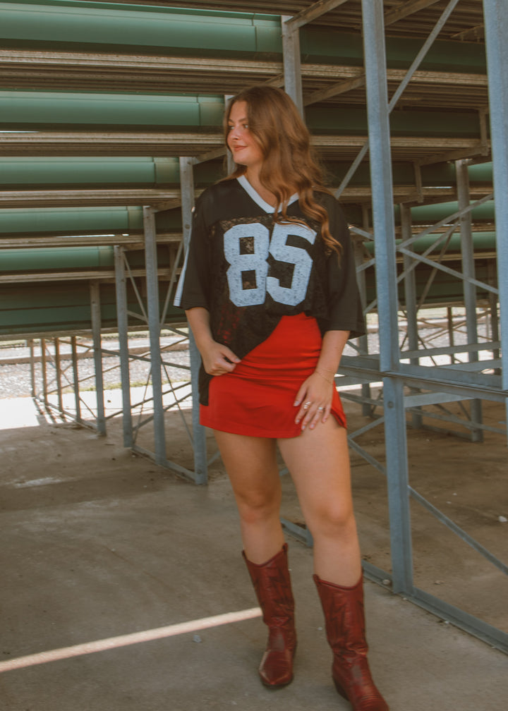 Woman wearing a sports jersey with number 85, red skirt, and brown boots in an indoor setting.
