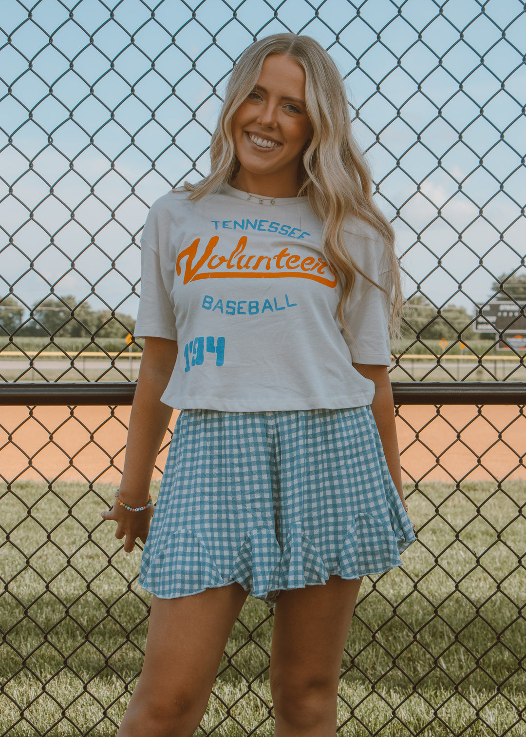 Woman wearing a 'Tennessee Volunteers Baseball' shirt and checkered shorts standing in front of a chain-link fence.