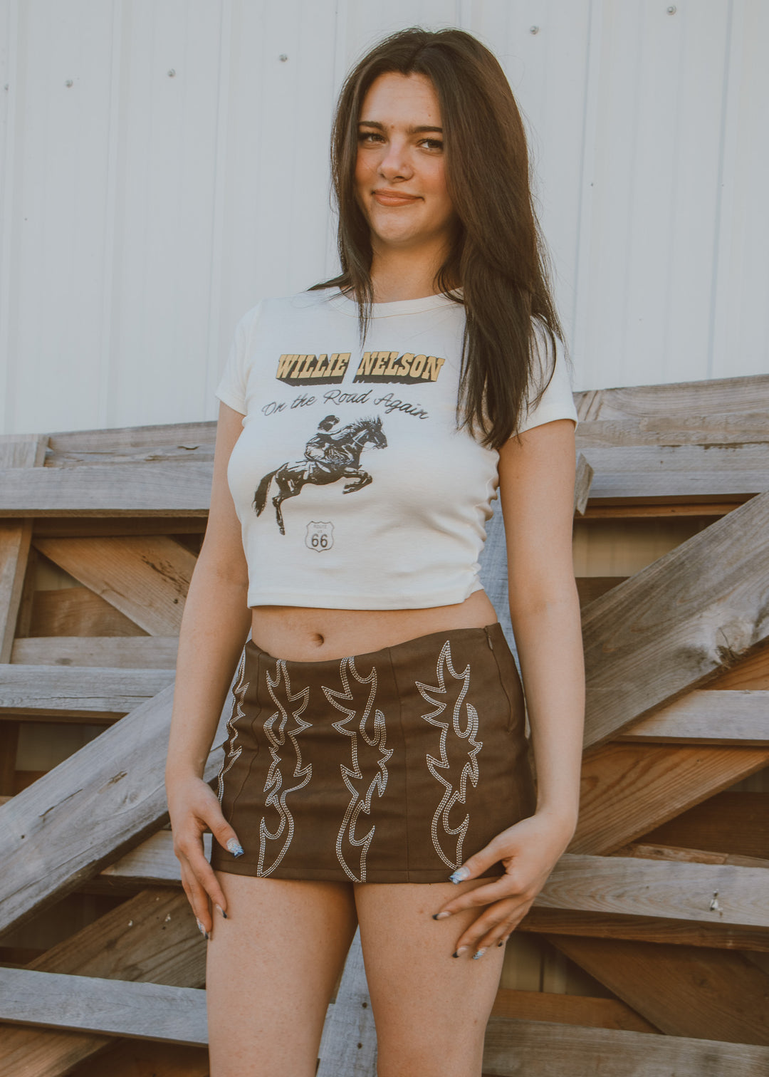 Woman wearing a white graphic t-shirt and brown skirts standing on wooden steps.