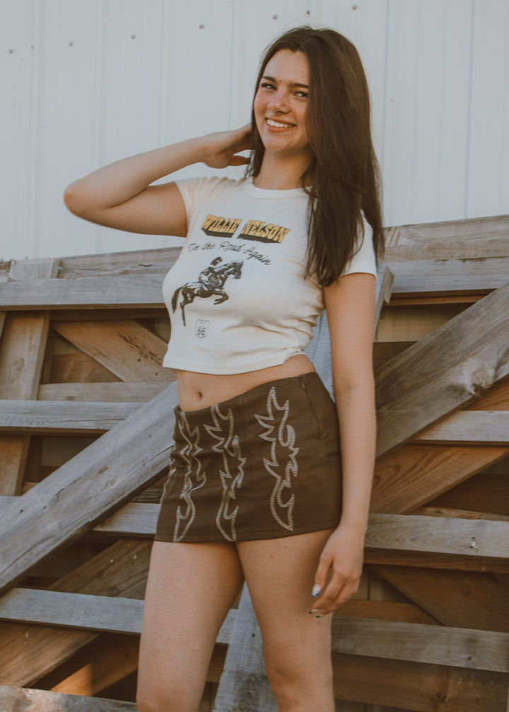 Woman wearing a graphic t-shirt and brown skirt standing against a wooden fence.