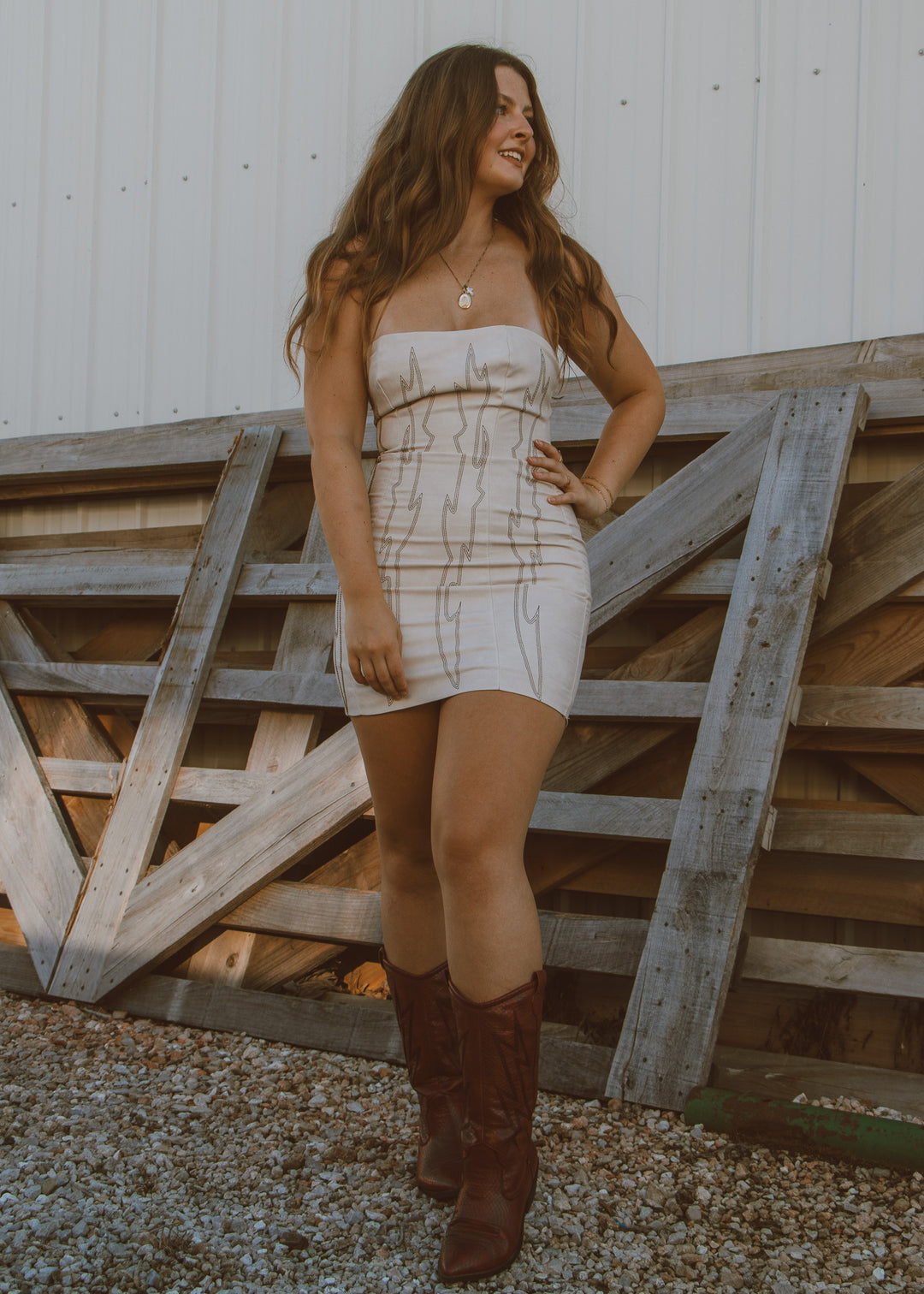 Woman in a strapless dress and brown boots standing in front of wooden gates.