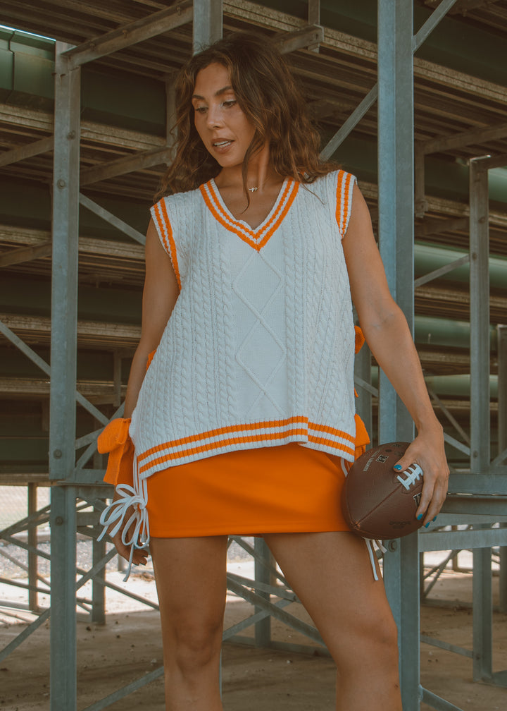 Woman in a white and orange dress holding a football, standing in front of metal beams.