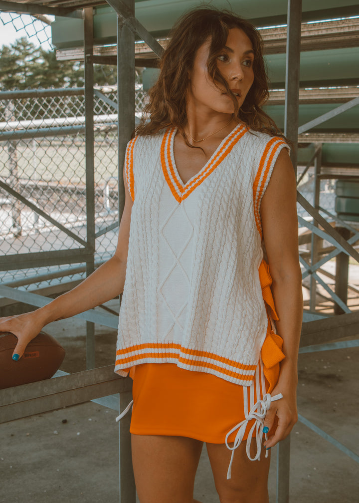 Woman wearing a white sleeveless top with orange accents and an orange skirt, standing outdoors.