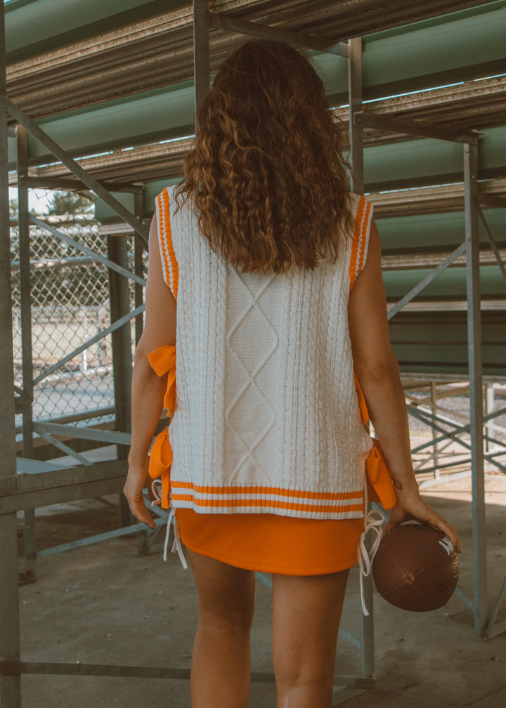 Woman in a white sleeveless top with orange trim and orange skirt holding a brown bag.