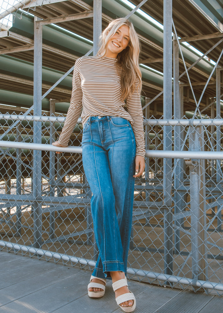Woman wearing a striped long-sleeve top and blue jeans standing in front of a metal fence.