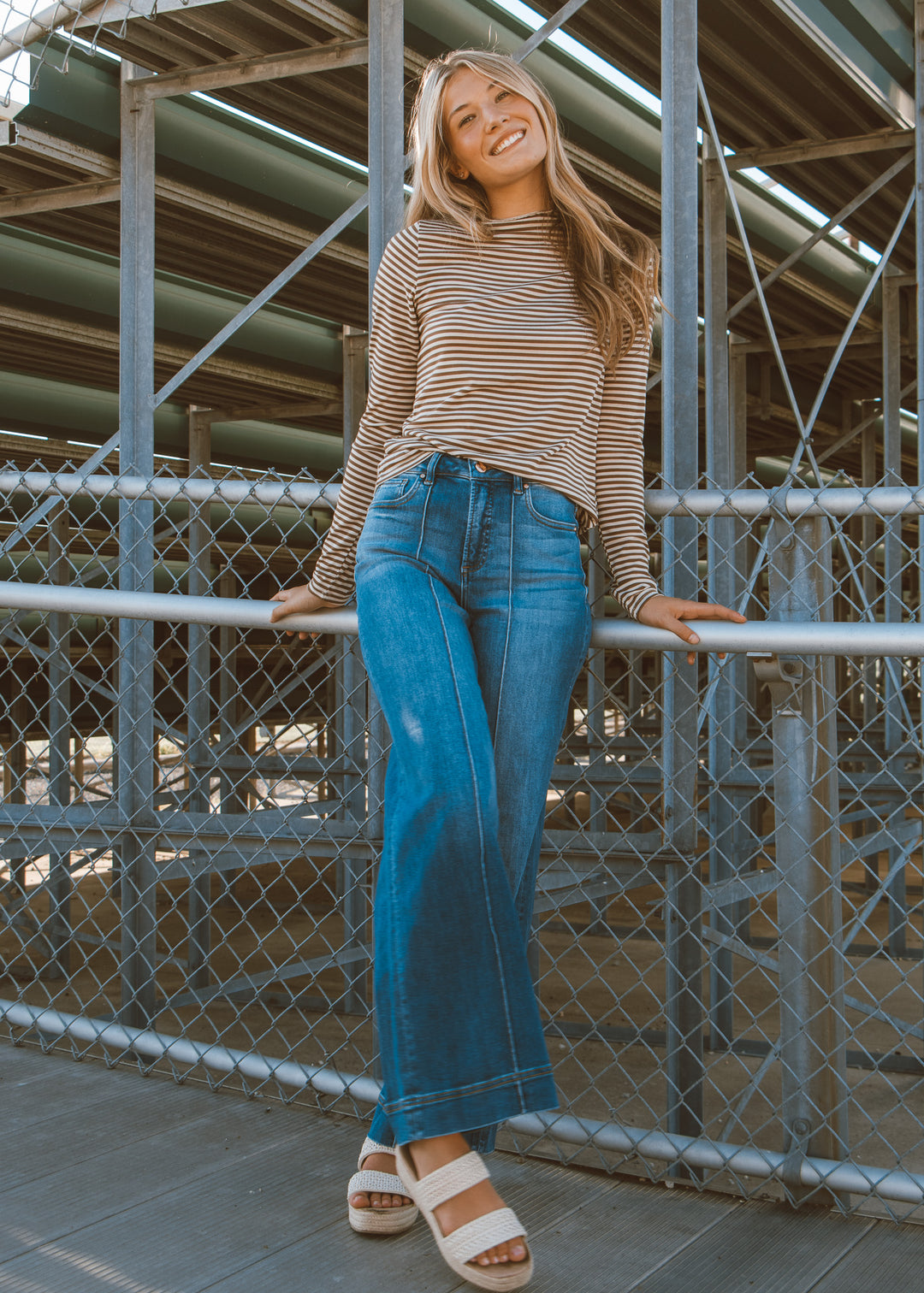Woman in striped shirt and blue jeans leaning against a metal railing.