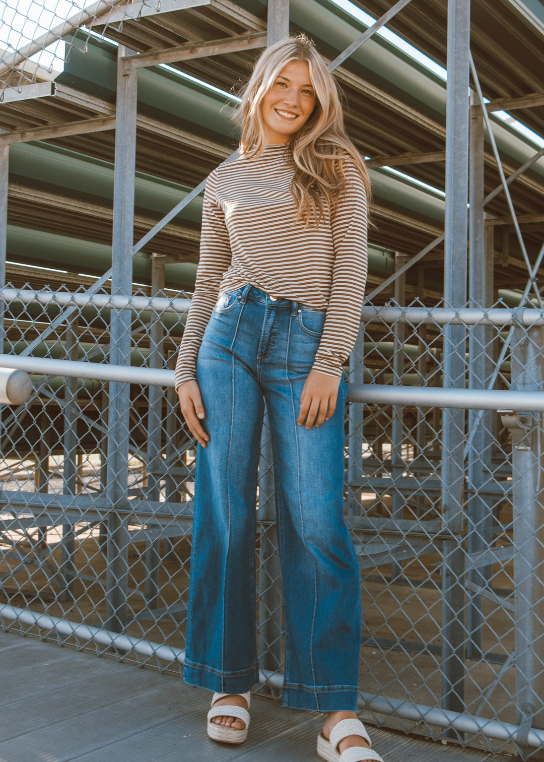 Woman wearing a striped sweater and blue jeans standing in front of a metal fence.