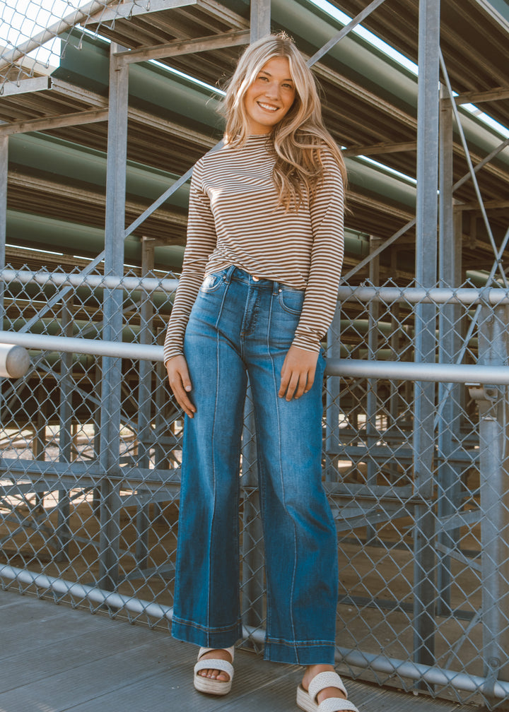 Woman wearing a striped sweater and blue jeans standing in front of a metal fence.