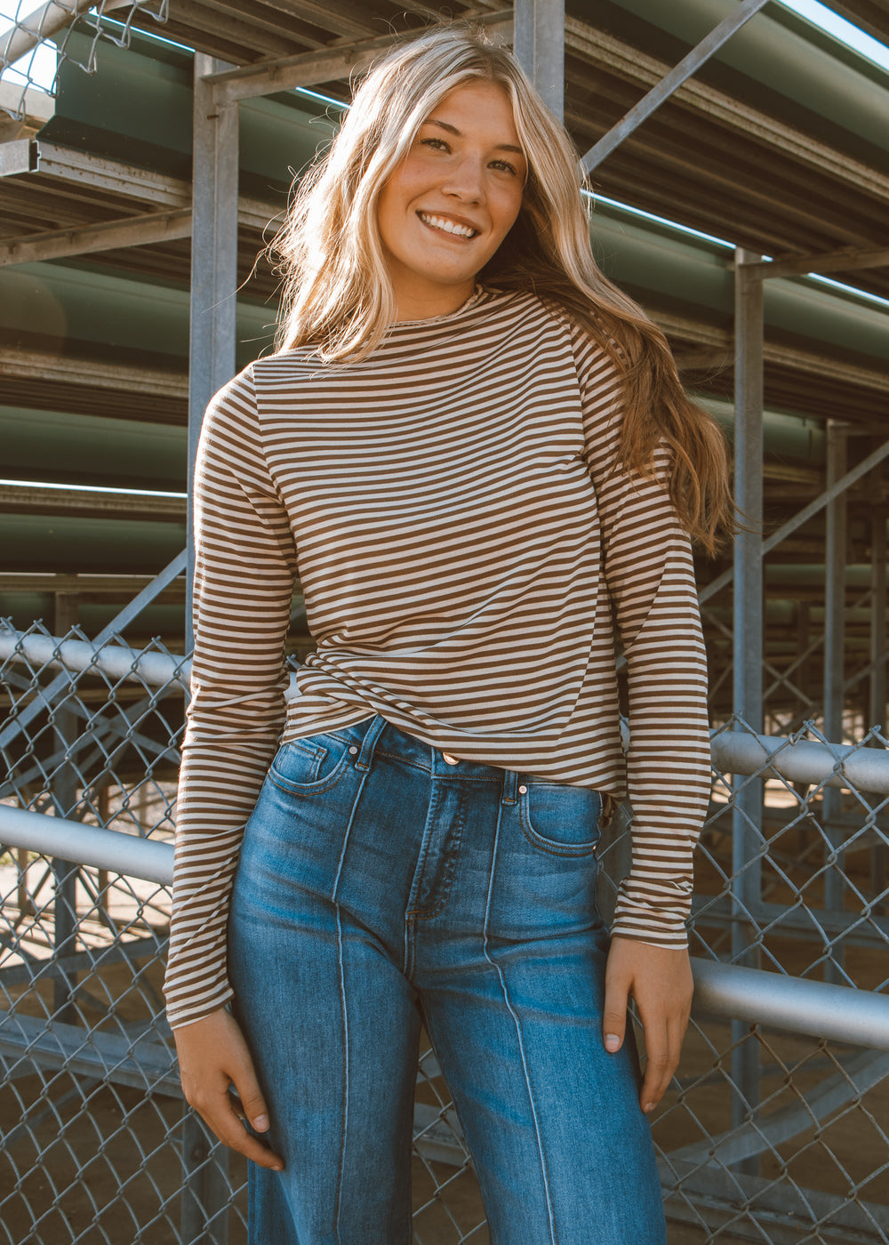 Woman wearing a striped shirt and blue jeans standing in front of a metal fence.