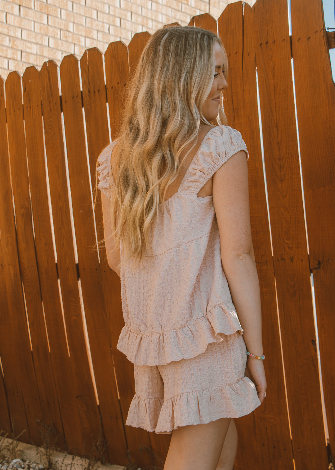 Woman wearing a light pink dress standing against a wooden fence.