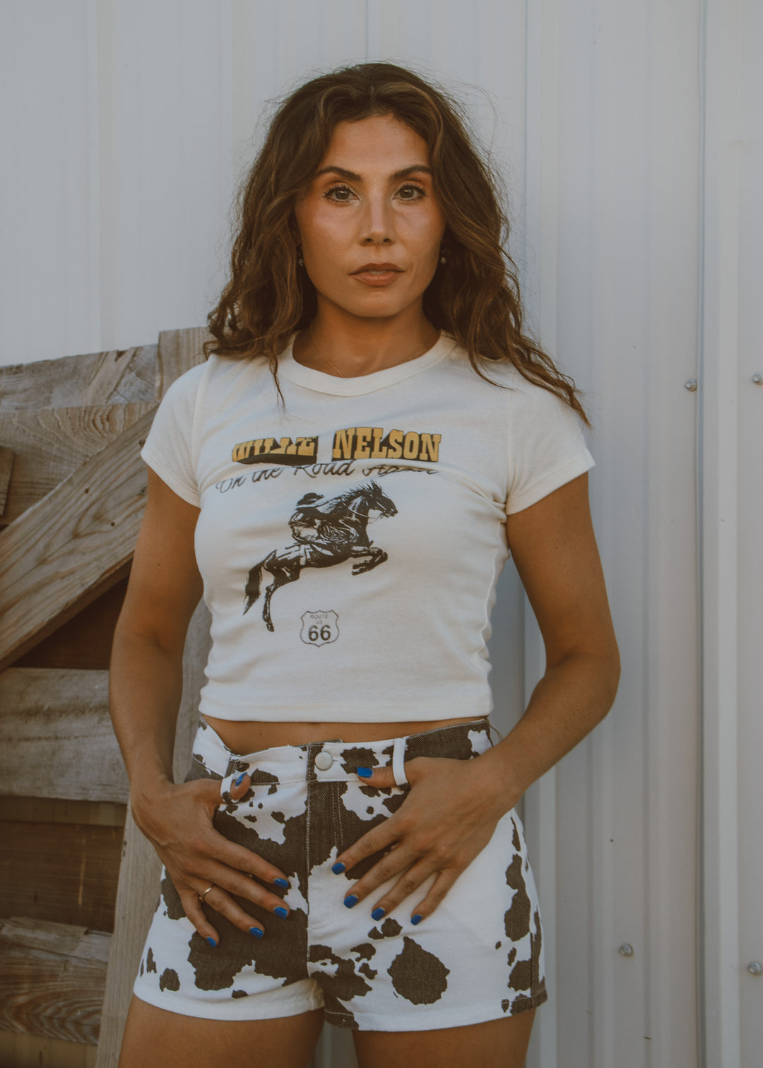 Woman wearing a graphic t-shirt and cow print shorts standing against a wooden fence.