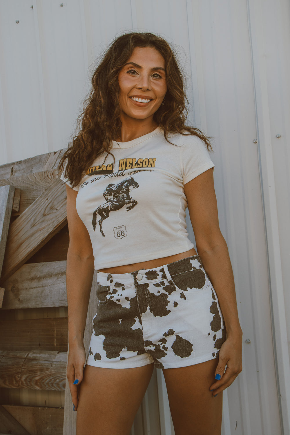 Woman wearing a white t-shirt with a graphic design and cow print shorts standing against a wooden background.