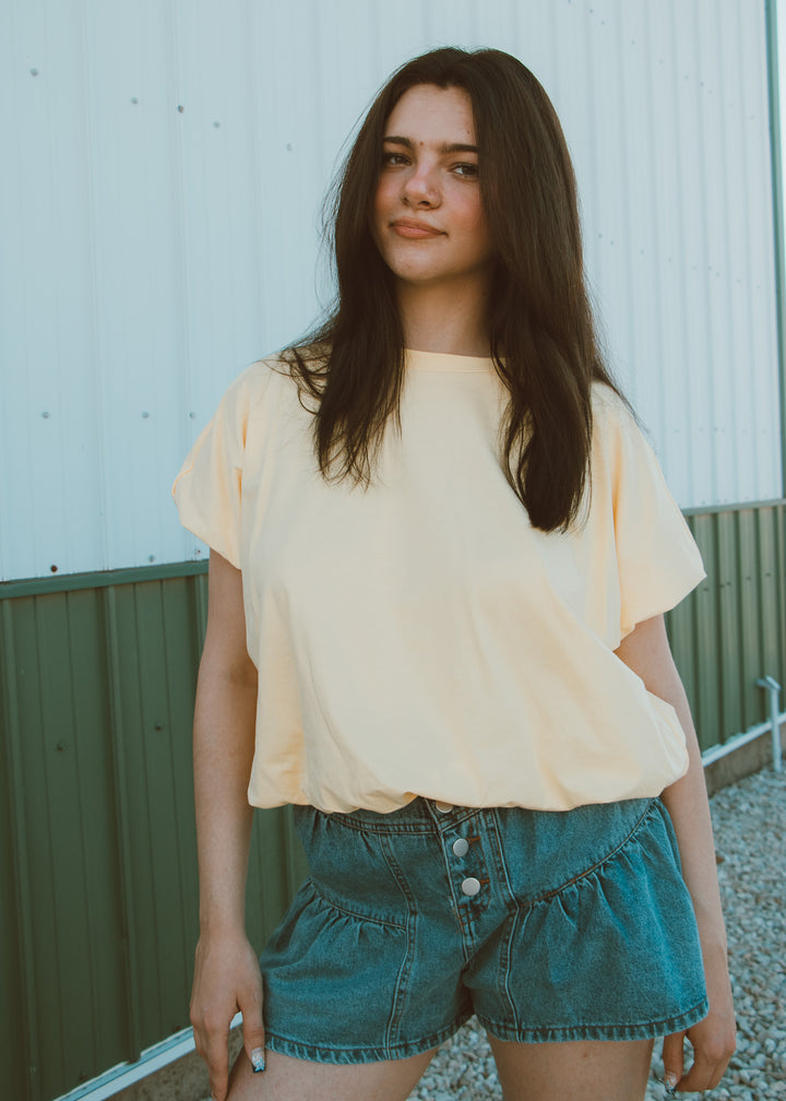 Woman wearing a yellow top and denim shorts standing against a light-colored wall.